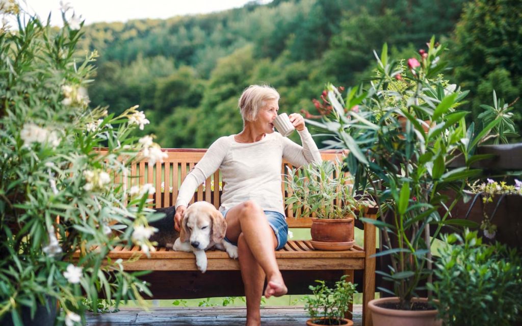 Frau sitzt mit Hund und einer Tasse Tee auf einer Bank im Garten :: Foto Adobe Stock